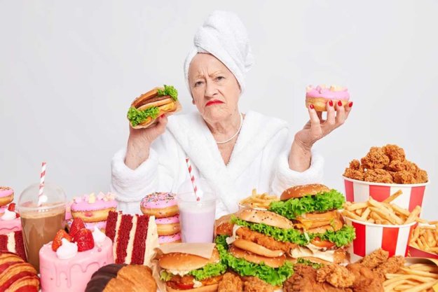 Displeased mature woman holds delicious doughnut and burger