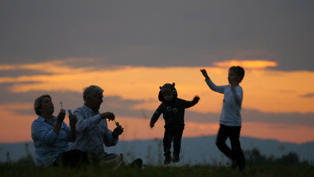 Grandparent sit on hill and blow bubbles, children playing, sund Grandparent sit on hill and blow bubbles, children playing, sundown sky