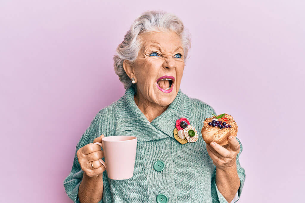 Senior grey-haired woman drinking a cup of coffee and eating bun