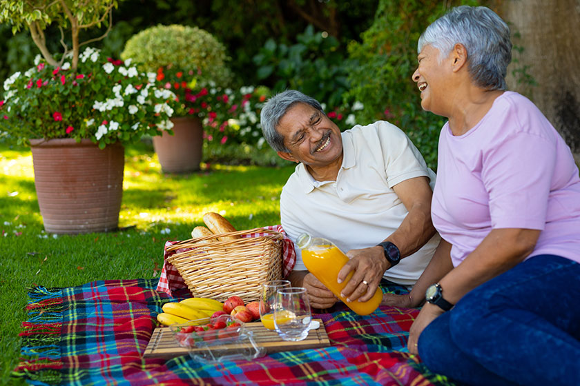 Smiling biracial senior man pouring juice in glass while enjoying picnic with wife in park. nature, happy, food, drink, unaltered, lifestyle, togetherness, love, picnic, enjoyment and retirement.
