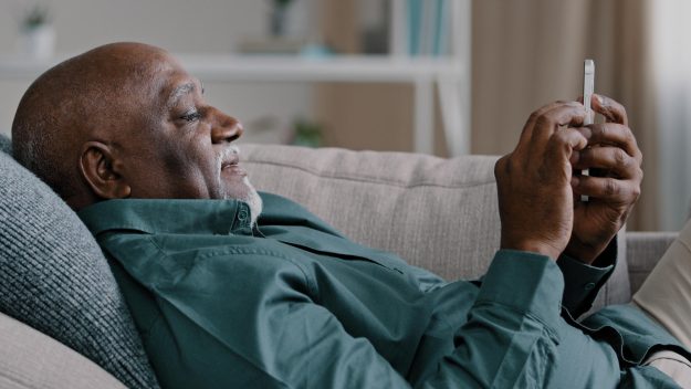 Relaxed old African American 60s bald bearded biracial man lie on soft comfortable couch at living room spend lazy weekend watching video mobile phone reading news calm elderly male rest use cellphone